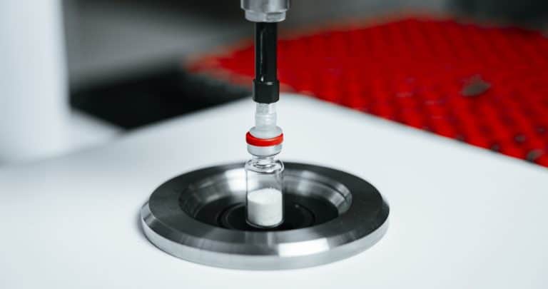Close-up of a vial being filled with white powder by a machine, with a red grip component visible, as part of the headspace analysis process in lab testing.