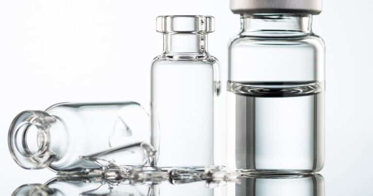 Three glass vials on a reflective surface, two empty and one filled with liquid and sealed with a red cap, scattered with small glass shards and droplets.
