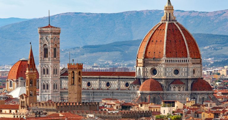 A panoramic view of Florence, Italy, featuring the Florence Cathedral with its iconic red dome and bell tower against a backdrop of mountains.