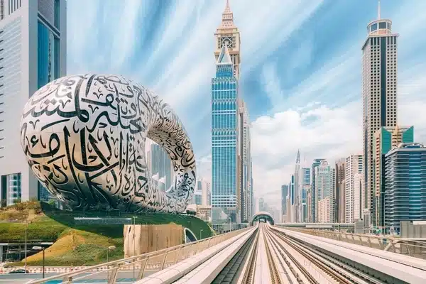 Futuristic cityscape featuring the Museum of the Future with Arabic calligraphy and multiple high-rise buildings under a blue sky. Foreground includes train tracks leading into the scene.