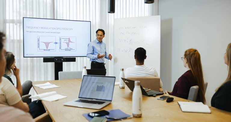 At the Lighthouse CCIT Academy, a man presents data on a screen and whiteboard to an engaged group seated at a conference table, their laptops and notebooks open and ready.