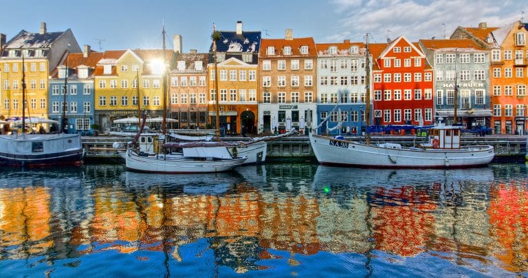 Colorful buildings and docked boats along a canal in Nyhavn, Copenhagen, reflect on the water under a clear blue sky, while a distant lighthouse adds charm to this picturesque scene.