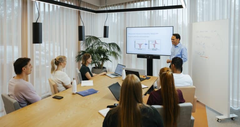 A man presents on spectroscopy to five people seated around a conference table with laptops, a whiteboard, and a screen displaying a graph related to the Container Closure Integrity Test.